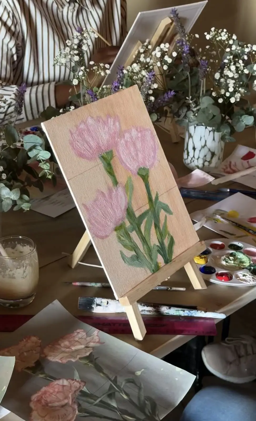 A canvas of pink tulips on a small easel beside a paint palette and fresh flowers